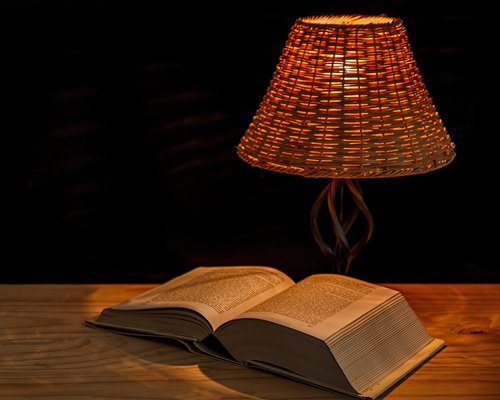 Person reading a printed book under a warm amber bedside lamp at night
